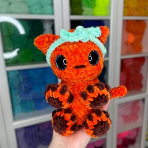 May include: A crocheted orange and brown cat toy with a light blue headband. The toy has large black eyes and a small, black nose.