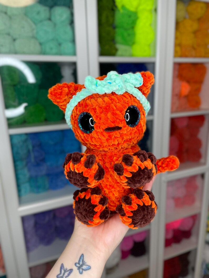 May include: A crocheted orange and brown cat toy with a light blue headband. The toy has large black eyes and a small, black nose.