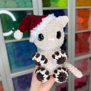 May include: A crocheted cat wearing a red and white Santa hat. The cat is beige with brown paw prints on its paws and tail.