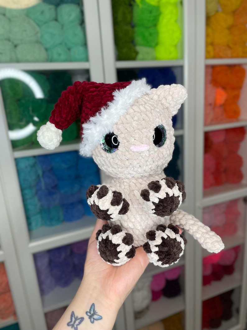 May include: A crocheted cat wearing a red and white Santa hat. The cat is beige with brown paw prints on its paws and tail.