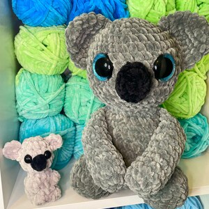 K&ouml;nnte beinhalten: Zwei geh&auml;kelte Koala-Pl&uuml;schtiere, eines grau und eines helllila, sitzen auf einem Regal mit Garnkn&auml;uel in den Farben Blau, Gr&uuml;n und Hellblau.