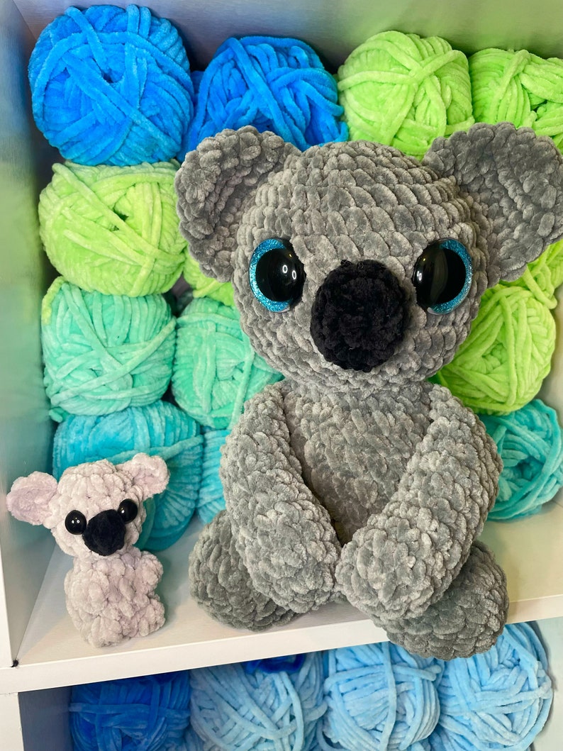 K&ouml;nnte beinhalten: Zwei geh&auml;kelte Koala-Pl&uuml;schtiere, eines grau und eines helllila, sitzen auf einem Regal mit Garnkn&auml;uel in den Farben Blau, Gr&uuml;n und Hellblau.