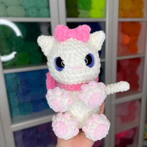 May include: White crocheted cat plush toy with pink bow, pink paws, and blue eyes. The toy is being held by a hand with a butterfly tattoo.