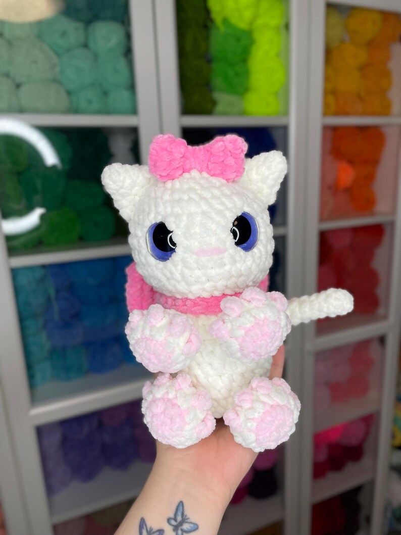 May include: White crocheted cat plush toy with pink bow, pink paws, and blue eyes. The toy is being held by a hand with a butterfly tattoo.