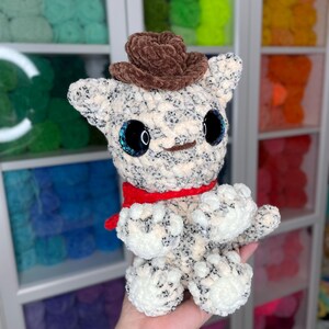 May include: A crocheted plush toy in the shape of a cat wearing a brown cowboy hat and a red scarf. The toy has large, black eyes with blue glitter accents.