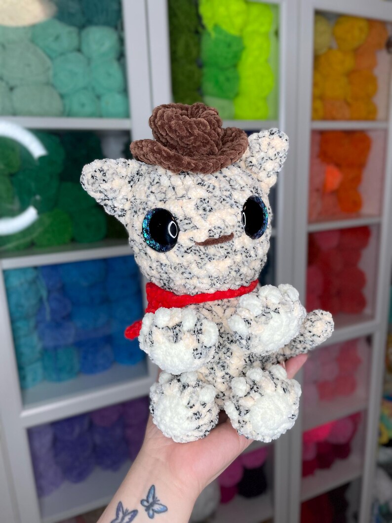May include: A crocheted plush toy in the shape of a cat wearing a brown cowboy hat and a red scarf. The toy has large, black eyes with blue glitter accents.