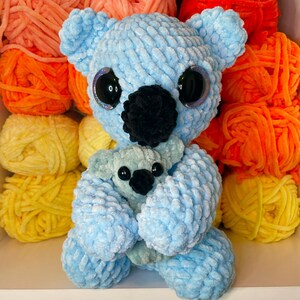K&ouml;nnte beinhalten: Ein hellblauer geh&auml;kelter Koala, der einen kleineren hellblauen geh&auml;kelten Koala h&auml;lt. Die Koalas haben schwarze Augen und Nasen. Die Koalas sitzen auf einem wei&szlig;en Regal mit bunten Wollkn&auml;ueln dahinter.
