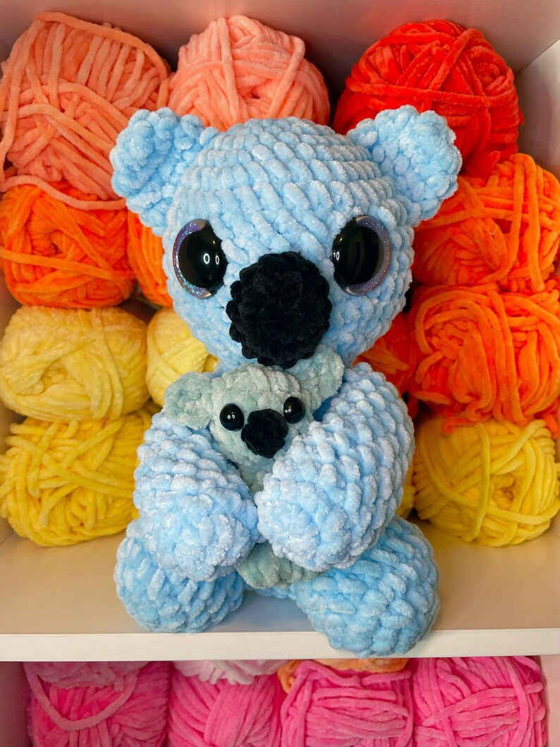 K&ouml;nnte beinhalten: Ein hellblauer geh&auml;kelter Koala, der einen kleineren hellblauen geh&auml;kelten Koala h&auml;lt. Die Koalas haben schwarze Augen und Nasen. Die Koalas sitzen auf einem wei&szlig;en Regal mit bunten Wollkn&auml;ueln dahinter.