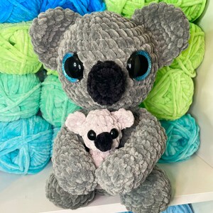K&ouml;nnte beinhalten: Ein grauer geh&auml;kelter Koala, der einen kleineren rosa geh&auml;kelten Koala h&auml;lt. Die Koalas haben schwarze Augen mit blauem Glitzer und schwarze Nasen. Die Koalas sind von Garnkn&auml;ueln in verschiedenen Blaut&ouml;nen und Gr&uuml;nt&ouml;nen umgeben.