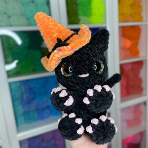 May include: A black crocheted cat wearing an orange and black witch hat with glitter. The cat has pink paw pads and sparkly eyes.