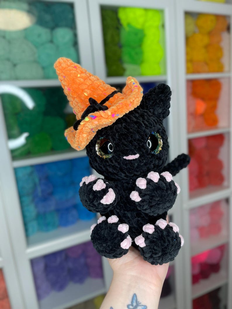 May include: A black crocheted cat wearing an orange and black witch hat with glitter. The cat has pink paw pads and sparkly eyes.