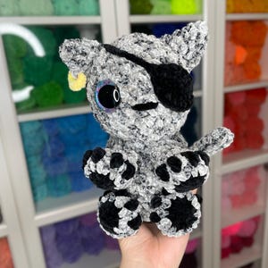 May include: A crocheted gray and black cat plush toy with a black eye patch and a yellow ear tag. The toy has large, black eyes with iridescent pupils.