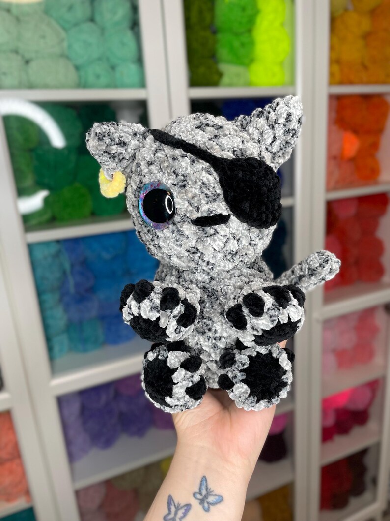 May include: A crocheted gray and black cat plush toy with a black eye patch and a yellow ear tag. The toy has large, black eyes with iridescent pupils.