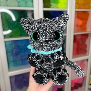 May include: A black and white crocheted plush toy cat with teal eyes and a light blue collar. The cat is sitting on a hand.