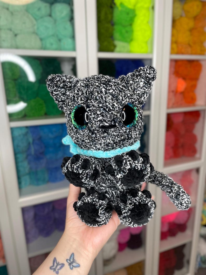 May include: A black and white crocheted plush toy cat with teal eyes and a light blue collar. The cat is sitting on a hand.