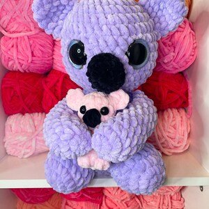 K&ouml;nnte beinhalten: Ein lila geh&auml;kelter Koala, der einen rosa geh&auml;kelten Baby-Koala h&auml;lt. Die Koalas sitzen auf einem Regal mit rosa, roten und orangen Garnkn&auml;ueln.