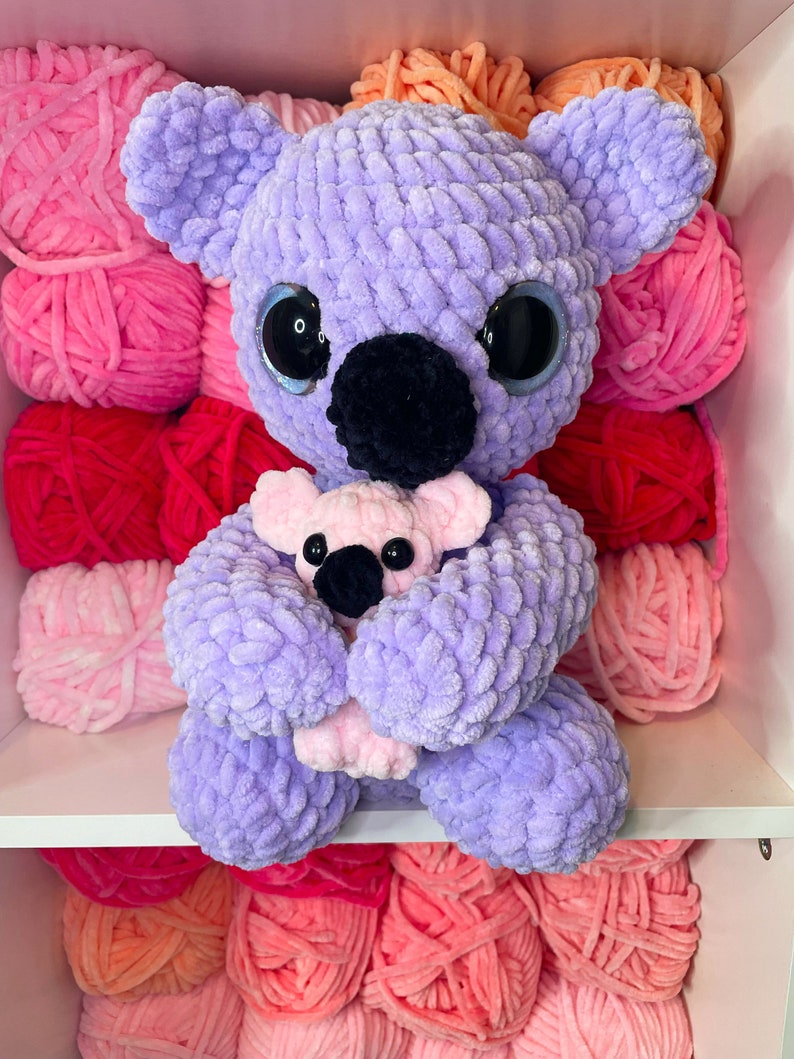 K&ouml;nnte beinhalten: Ein lila geh&auml;kelter Koala, der einen rosa geh&auml;kelten Baby-Koala h&auml;lt. Die Koalas sitzen auf einem Regal mit rosa, roten und orangen Garnkn&auml;ueln.
