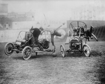 May include: Two vintage cars are racing on a grassy field. One car is being driven by a man who is leaning out of the car and kicking a ball. The other car is being driven by a man who is holding onto the steering wheel. The cars are both in motion and the ball is in the air.
