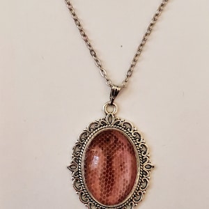 May include: Silver-toned necklace with an oval pendant. The pendant features a reddish-brown, textured design within an ornate, filigree frame. The chain is delicate and hangs from the pendant.