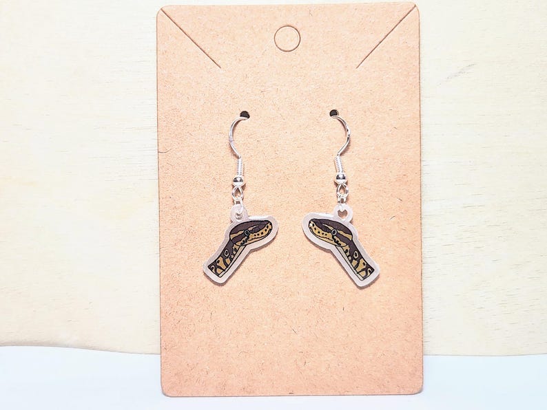 Może przedstawiać: Para kolczyk&oacute;w z motywem głowy węża. Kolczyki mają srebrne haczyki i przezroczyste, prostokątne zawieszki z brązowo-ż&oacute;łtymi ilustracjami głowy węża. Są prezentowane na brązowej karcie.