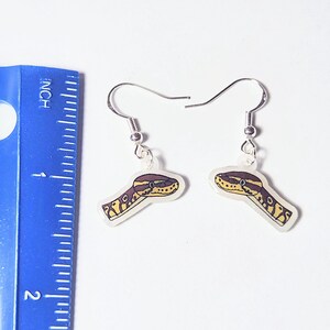 Może przedstawiać: Para srebrnych kolczyk&oacute;w z motywem głowy węża. Głowy węża są ż&oacute;łte, brązowe i białe, z przezroczystym tłem. Niebieska linijka znajduje się w tle.