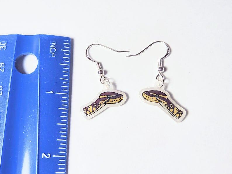 Może przedstawiać: Para srebrnych kolczyk&oacute;w z motywem głowy węża. Głowy węża są ż&oacute;łte, brązowe i białe, z przezroczystym tłem. Niebieska linijka znajduje się w tle.