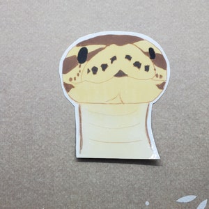 May include: A sticker featuring a cartoon illustration of a snake's head. The snake has a tan and brown pattern with black eyes and a light yellow face. The sticker is on a brown background.