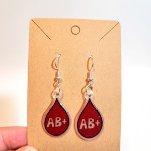 May include: A pair of dangle earrings featuring a blood drop design with the text "AB+" in white. The earrings have a clear outline and silver-colored hooks, displayed on a brown card.