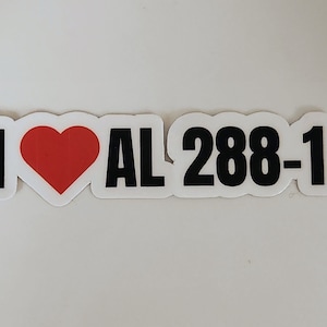May include: A white sticker with a black border. The sticker features the phrase "I ♥ AL 288-1". The heart is red. The text "AL 288-1" is in a bold, sans-serif font.