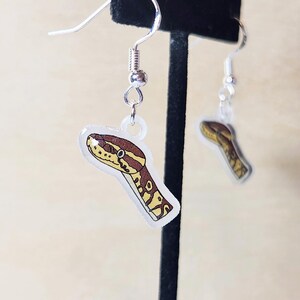 Może przedstawiać: Kolczyki wiszące z motywem głowy węża. Kolczyki mają przezroczyste, błyszczące wykończenie ze srebrnymi haczykami. Głowa węża jest brązowa, beżowa i ż&oacute;łta, ze szczeg&oacute;łowym wzorem. Kolczyki są prezentowane na neutralnym tle.