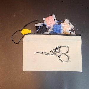 May include: A cream-colored zippered pouch with a black scissor design and a black zipper. The pouch is filled with spools of colorful embroidery floss in pink, blue, and cream. A yellow whistle is attached to the zipper.