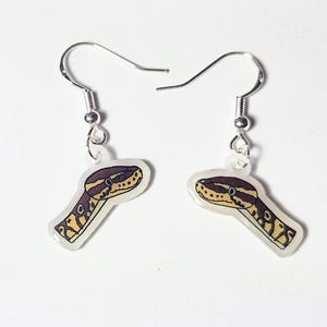May include: A pair of dangle earrings featuring a snake head design. The earrings have a clear background with a silver hook and a detailed illustration of a snake head in shades of brown, yellow, and black.