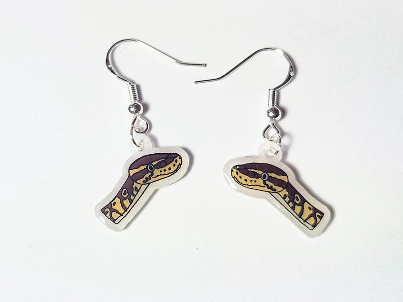 Może przedstawiać: Para kolczyk&oacute;w z motywem głowy węża. Kolczyki mają przezroczyste tło ze srebrnym haczykiem i szczeg&oacute;łową ilustracją głowy węża w odcieniach brązu, ż&oacute;łci i czerni.