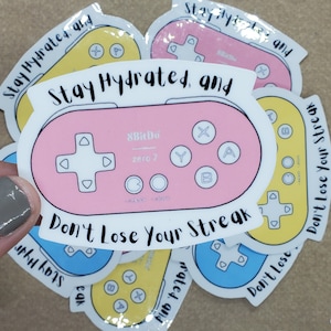 May include: A collection of colorful stickers featuring a video game controller design. The central sticker is pink with white text and the words "Stay Hydrated, and Don't Lose Your Streak." Other stickers are yellow and blue.
