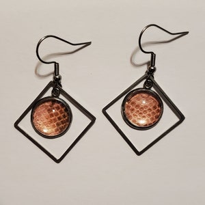 Może przedstawiać: Para kolczyków wiszących. Każdy kolczyk ma ciemnoszarą, kwadratową ramkę z okrągłym, brzoskwiniowym środkiem. Środek ma wzór skóry węża. Kolczyki mają zapięcie na haczyk.