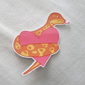 May include: A sticker featuring a cartoon snake wrapped around a pink heart. The snake is orange with yellow spots and a yellow head. The heart is a bright pink color with a white border. The snake's tongue is sticking out.