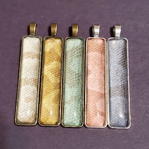 May include: Five rectangular pendants with snake skin patterns in various colors: white, gold, green, pink, and gray. Each pendant has a metal frame and a loop at the top for a chain. The pendants are displayed on a purple surface.