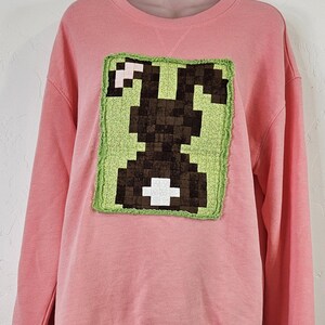 May include: A long-sleeve crewneck sweatshirt in a solid light pink color. The front features a rectangular fabric appliqué with a pixelated dark brown rabbit silhouette on a light green background, finished with a frayed, stitched edge.