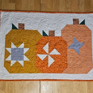 May include: A quilted wall hanging featuring three patchwork pumpkins in shades of orange, yellow, and brown. The pumpkins are decorated with white and gray stars and a white pinwheel design.