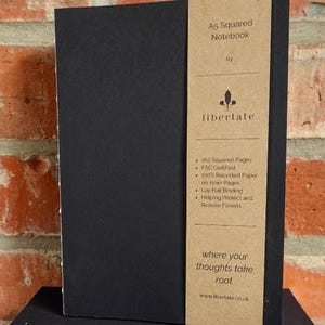May include: A5 squared notebook with a black cover and a brown paper band. The band features the text "A5 Squared Notebook" and "libertate". The notebook has 160 squared pages and is FSC certified.