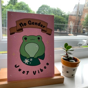 Puede incluir: Una tarjeta rosa con una pancarta marrón que dice "No Gender Just Vibes". Una rana verde con ojos grandes se sienta en un nenúfar con una flor blanca. La tarjeta está en un soporte de madera.