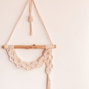May include: A macrame wall hanging in cream color. The design features a wooden dowel, a decorative knotted pattern, and a hanging tassel. The piece is suspended from a triangular macrame structure, with a small crystal accent.