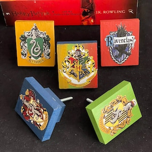 May include: Set of five wooden Harry Potter house crest knobs. The crests are painted in the colors of each house: Gryffindor (red and gold), Hufflepuff (yellow and black), Slytherin (green and silver), Ravenclaw (blue and bronze), and  a fifth crest with the words "Dormiens Nunquam", "Draco", and "Itillandus" in Latin.