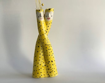 Puede incluir: Un jarrón de cerámica amarillo con lunares negros en forma de dos figuras entrelazadas con caras. El jarrón tiene una abertura estrecha en la parte superior y está decorado con detalles pintados, incluidos ojos, labios y cejas.