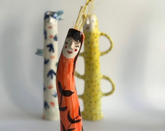 Puede incluir: Tres jarrones de cerámica con caras y patrones pintados. El jarrón del centro es naranja con diseños florales negros y la cara de una mujer. El jarrón de la izquierda es azul y blanco con un patrón geométrico. El jarrón de la derecha es amarillo con lunares negros.