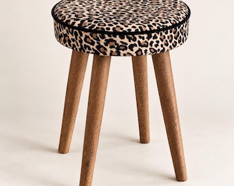 May include: A round stool featuring a cushioned seat upholstered in a tan, black, and brown leopard print fabric with black piping along the edge. The stool is supported by four tapered, light brown wooden legs with a visible natural wood grain.
