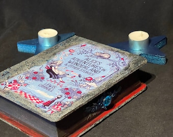 May include: A blue book with a red and white checkered pattern on the cover. The book is titled "Alice's Adventures in Wonderland & Other Stories" by Lewis Carroll. The book is decorated with a blue and silver star-shaped candle holder on each side.