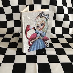 Op de afbeelding: Een wit dagboek met een textuur-omslag met een cartoonillustratie van Alice in Wonderland die een roze flamingo vasthoudt.