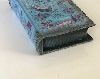 Puede incluir: Una caja gris en forma de libro con una cubierta azul que presenta una ilustración de Alicia en el País de las Maravillas. El título dice "Alice's Adventures in Wonderland & Other Stories" de Lewis Carroll.