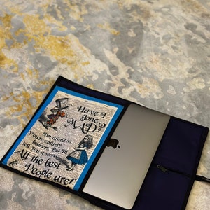May include: A blue and white laptop sleeve with a zipper closure. The front of the sleeve features a vintage book page with the text "Have I gone MAD?" and an illustration of Alice in Wonderland.